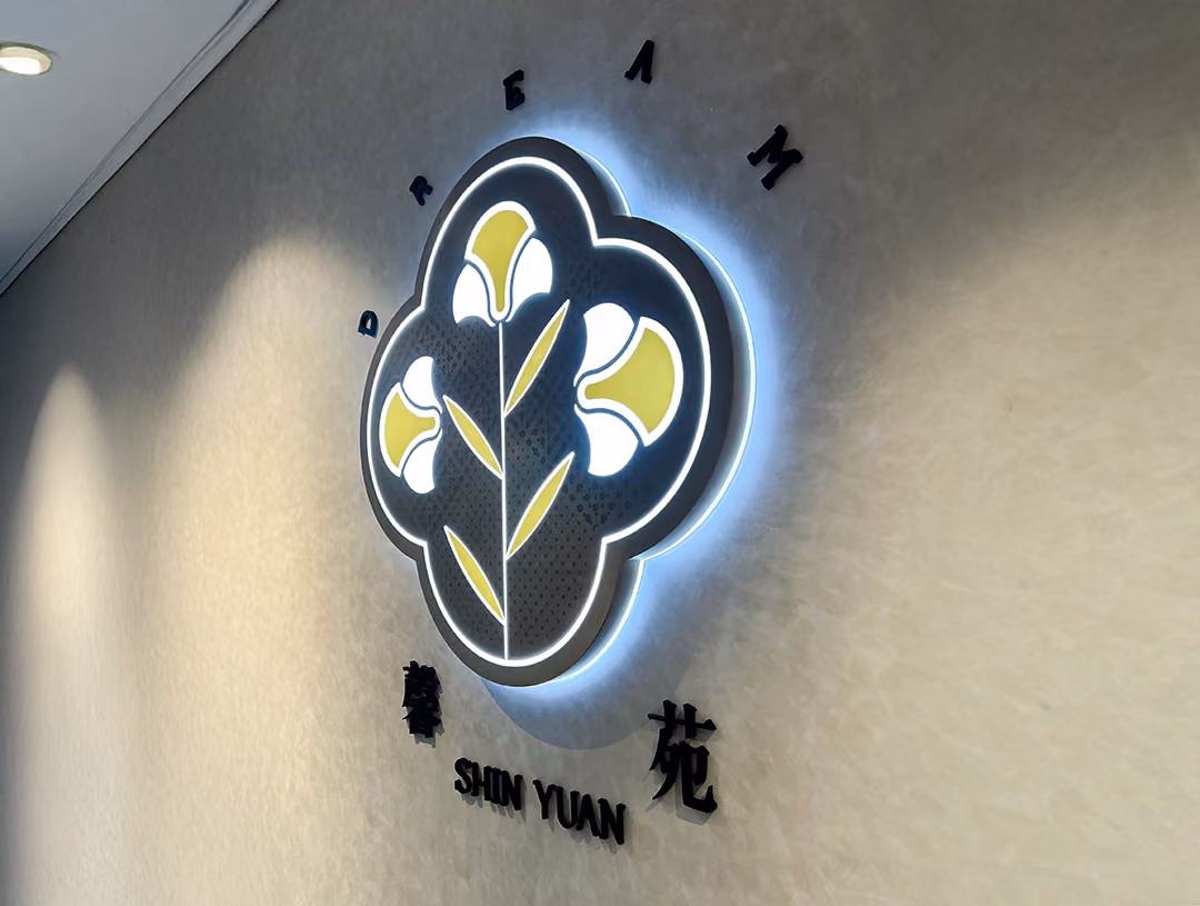 米白紋理牆上的深灰花形標誌板已點亮,邊緣透出冷白光暈,下方為黑色立體字