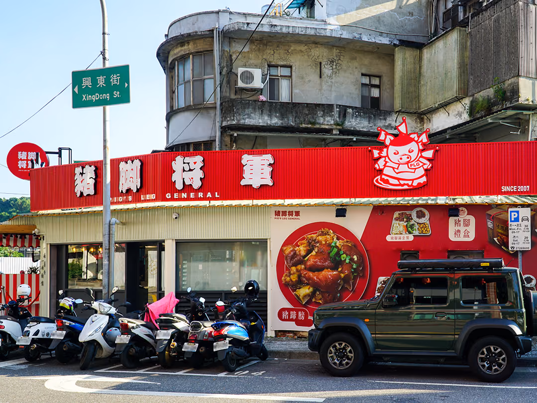 街角店面橫式紅色浪板招牌,白色立體字,右端紅白豬造型立體物凸出招牌上緣,下方牆面印滷豬腳大圖