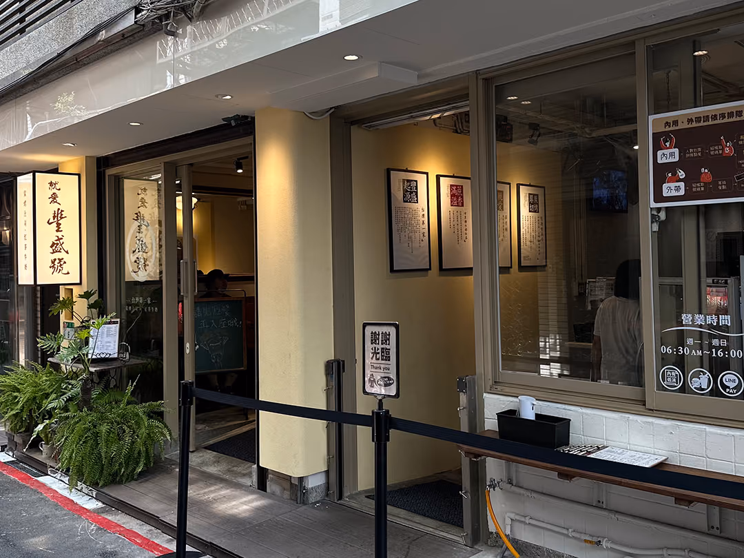 日間街邊店面,米黃立面配大片開窗,左側懸挑深框米白燈箱招牌,窗內暖黃燈光映著牆上四幅框裱書法