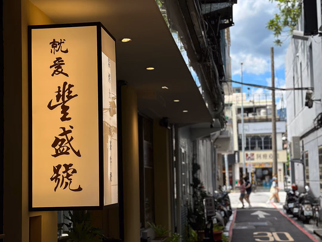 仰角看懸挑雙面燈箱,深色細框內米白燈面透出暖光,直書毛筆店名,背景藍天與街邊機車