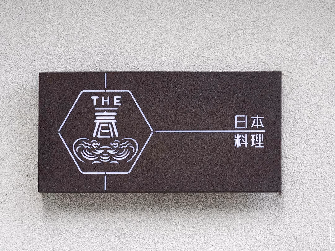 深褐色霧面箱型招牌特寫,白色細線六角圖記與日文字樣,粗紋白牆上投出淺淺陰影