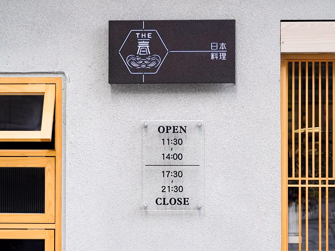 粗紋白牆上深褐色方形招牌,下方透明壓克力營業時間牌以金屬螺絲固定,左木框窗、右木格柵門,日間未點燈