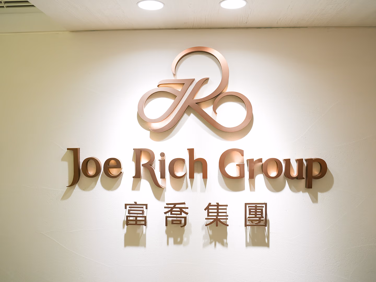 米白牆面上的玫瑰金金屬立體字,繞圈標誌置於中英文字之上,嵌燈斜照使字身投下柔和陰影